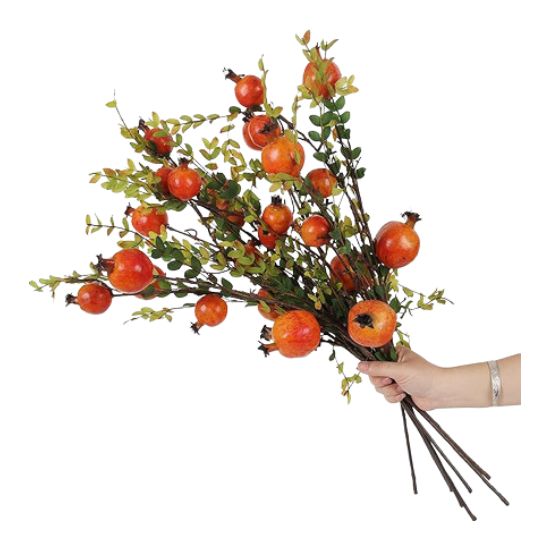 A hand holds a bouquet of artificial branches with green leaves and bright orange pomegranate fruits, perfect for learning how to decorate a footed bowl against a crisp white background.
