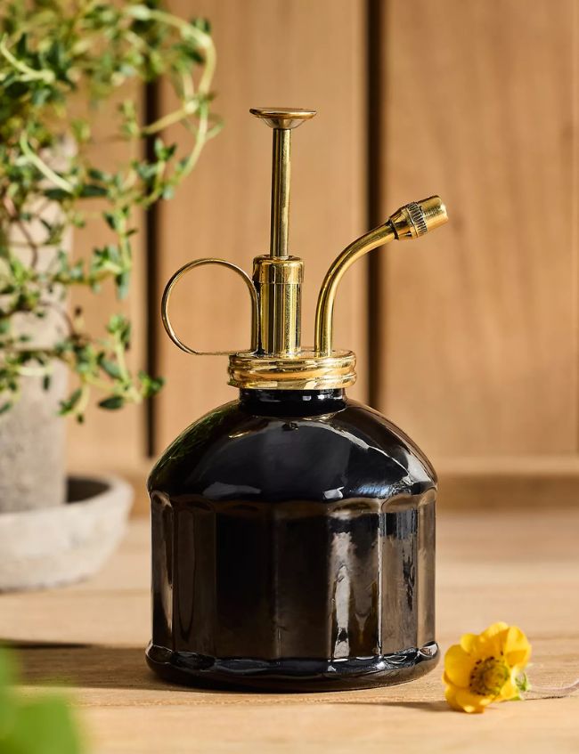 A small, elegant glass plant mister with a gold metal pump top sits on a wooden surface near a potted plant and yellow flower—one of our Best Picks for style and function, perfect to discover during the Terrain Sale.