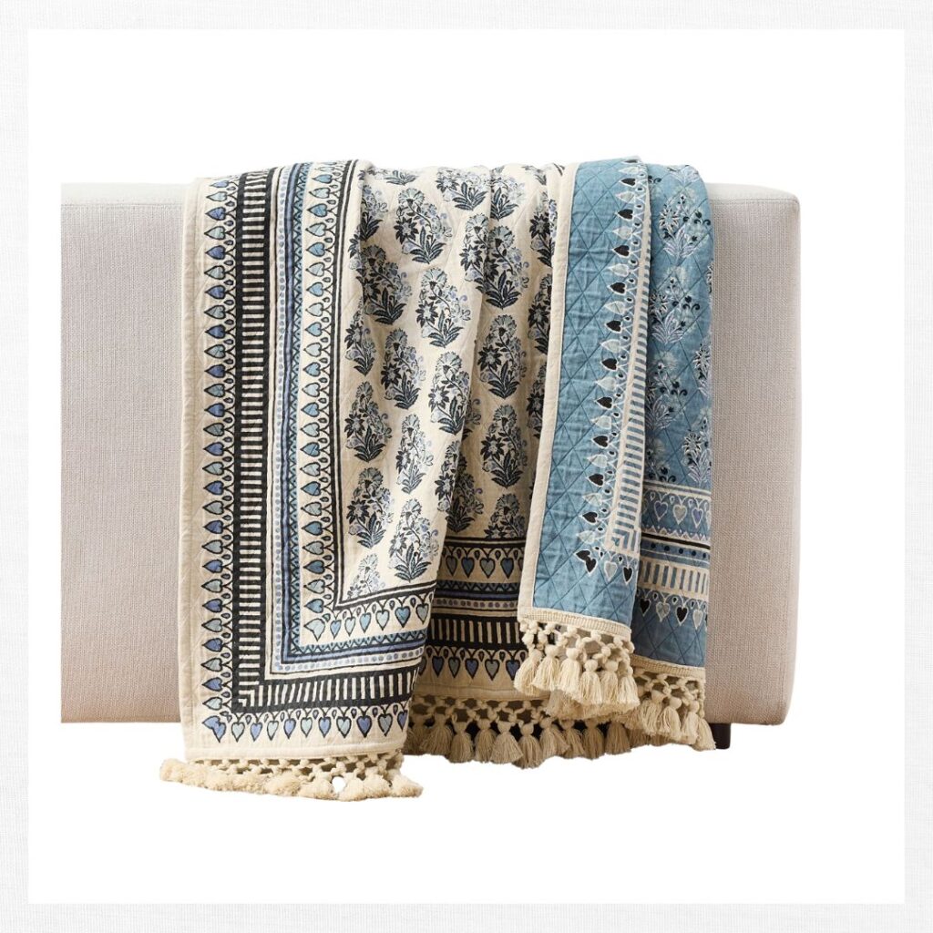 A patterned throw blanket with blue, black, and cream designs, featuring floral motifs and geometric borders, draped over a light-colored upholstered bench. The blanket has decorative tassels along the edges.