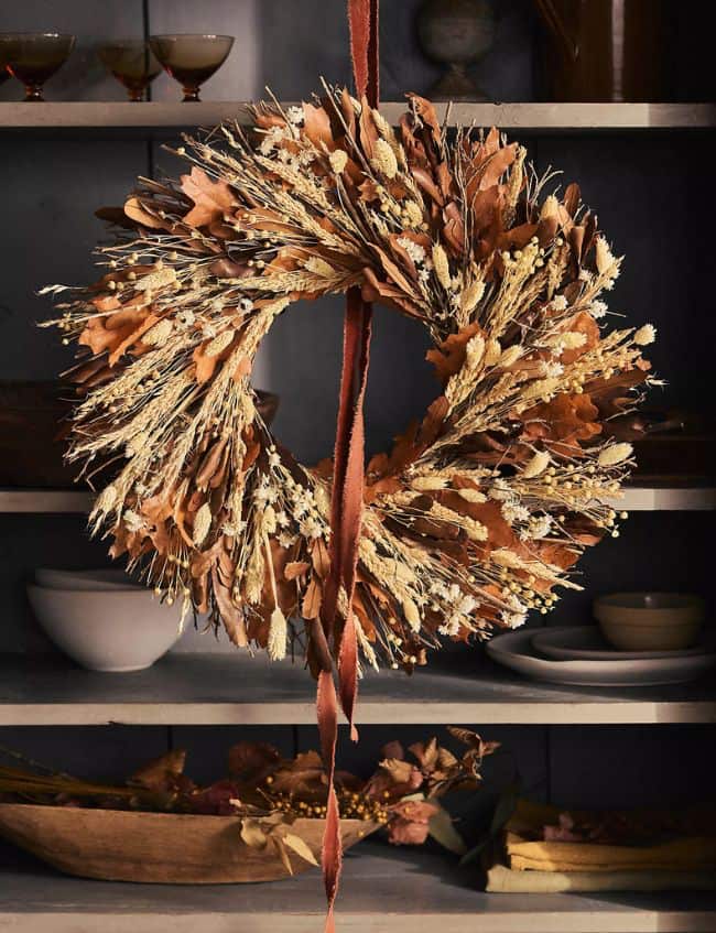 A rustic autumn wreath made of dried leaves, wheat, and grasses hangs by a brown ribbon in front of shelves with bowls and glasses. Featuring warm brown and gold tones, it’s one of our Top Picks for creating a cozy fall atmosphere during the Terrain Sale.