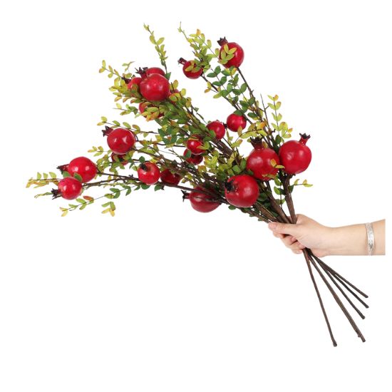A hand holds several artificial branches with red pomegranate fruits and green leaves, perfect for learning how to decorate a footed bowl, isolated on a white background.