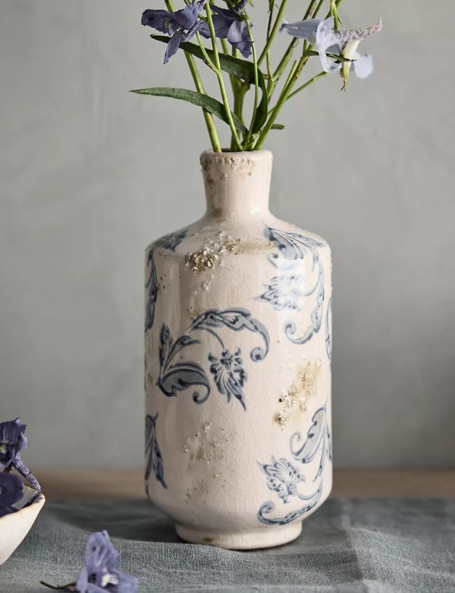 A cream-colored ceramic vase with blue floral patterns and a distressed finish holds a few purple flowers against a gray background—one of our Top Picks for the Terrain Sale.