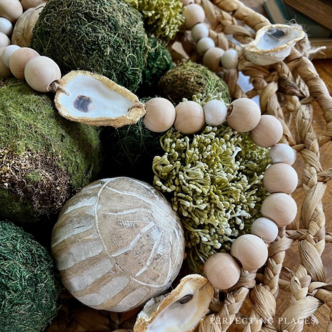 A close-up of a decorative arrangement featuring green moss balls, a textured white ornament, and a string of wooden beads with oyster shells—some of my Favorite Things. Beautiful details stand out against the woven basket background.