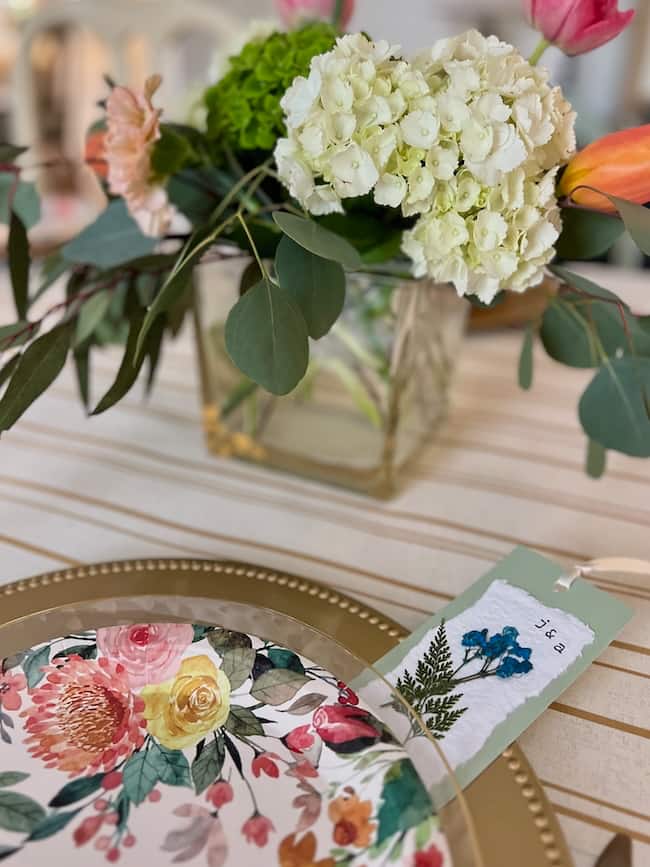 A decorative table setting with a gold charger plate, a floral-patterned plate, a floral arrangement in a glass vase, and a place card with pressed flowers and the name "Jessica" written on it.