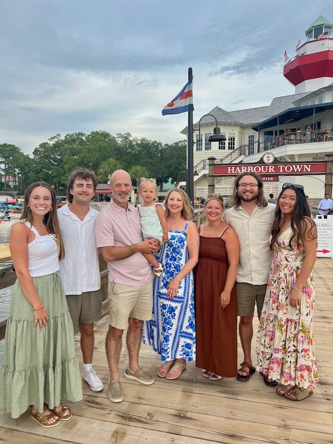 A group of eight people, including a child, stand smiling on a wooden dock in front of the Harbour Town pier. They are dressed in casual summer clothes, with a lighthouse and buildings visible in the background.