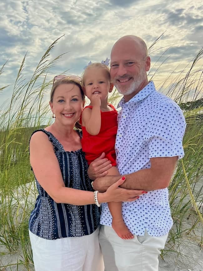 A smiling couple stands on a beach holding a young girl in a red dress. Tall grass surrounds them, and the sky is partly cloudy in the background.