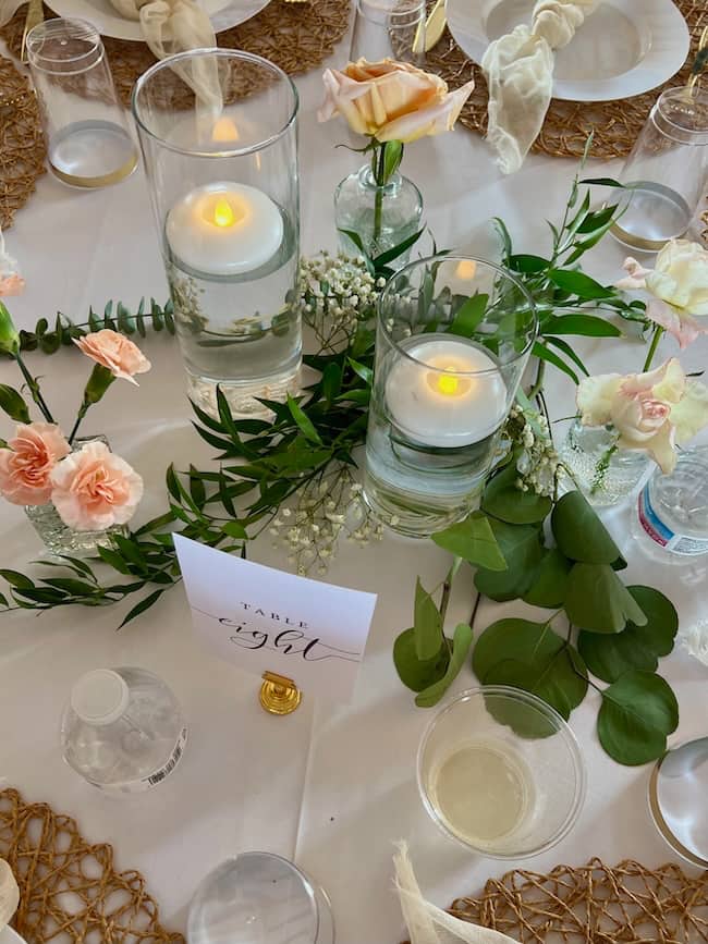A wedding table centerpiece with floating candles in glass vases, blush roses, greenery, and a card reading “Table Eight.” Water bottles, clear cups, and woven placemats are also visible on the table.