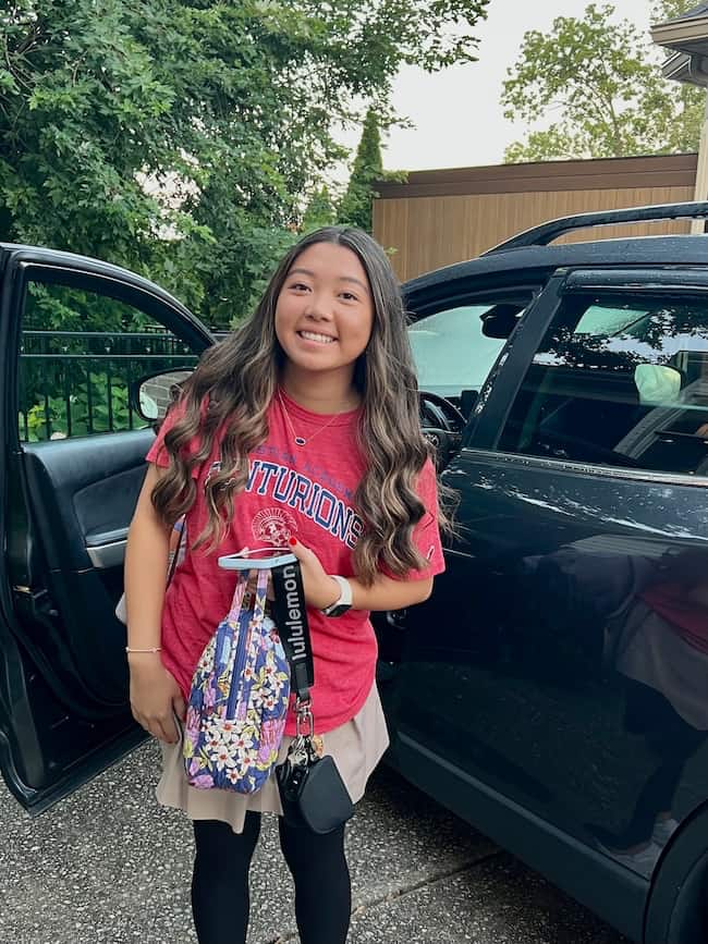 A smiling young woman with long hair stands beside an open car door, wearing a red "Centurions" shirt and holding a phone, a patterned face mask, and a lanyard. Green trees and a building are in the background.