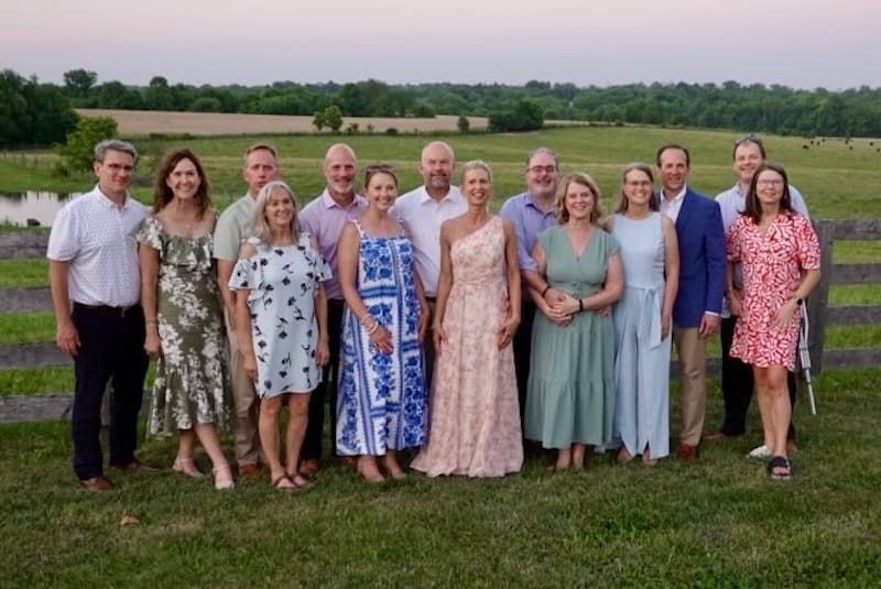 A group of fourteen adults poses together outside in front of a wooden fence, with green fields and trees in the background at sunset. They are dressed in summer clothes and smiling at the camera.