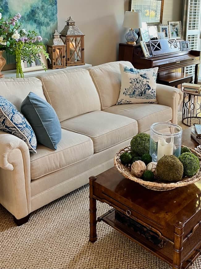 Beige sofa with blue and patterned pillows in a cozy living room; wooden coffee table with a decorative tray, moss balls, and candles atop textuneutral textured area rugs; piano, lanterns, and artwork in the background.