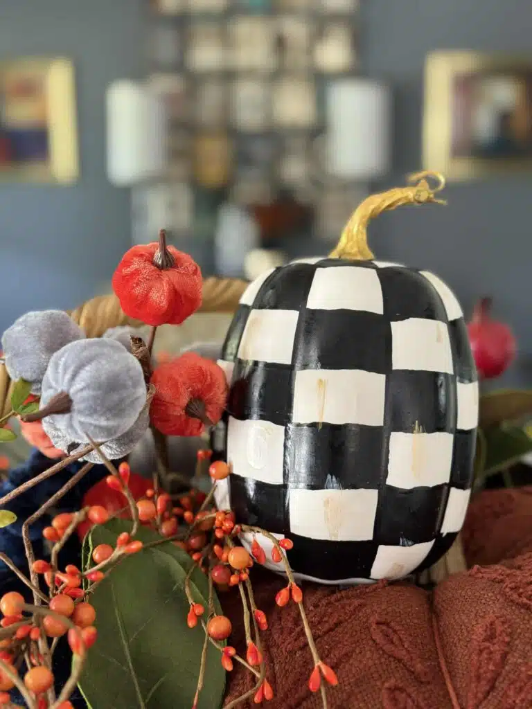 A decorative black-and-white checkered pumpkin sits among artificial red, purple, and orange berries and leaves, creating a festive autumn centerpiece—perfect for some cozy weekend or Saturday inspiration. The background is softly blurred.