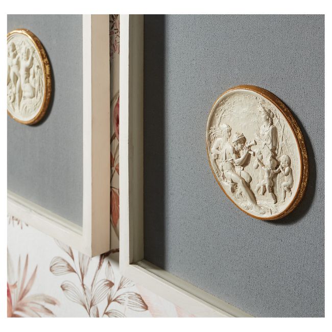 A close-up of two framed circular relief artworks, created using DIY intaglio art techniques, with classical figures, mounted on a textured gray wall above wallpaper with pink and brown floral patterns.