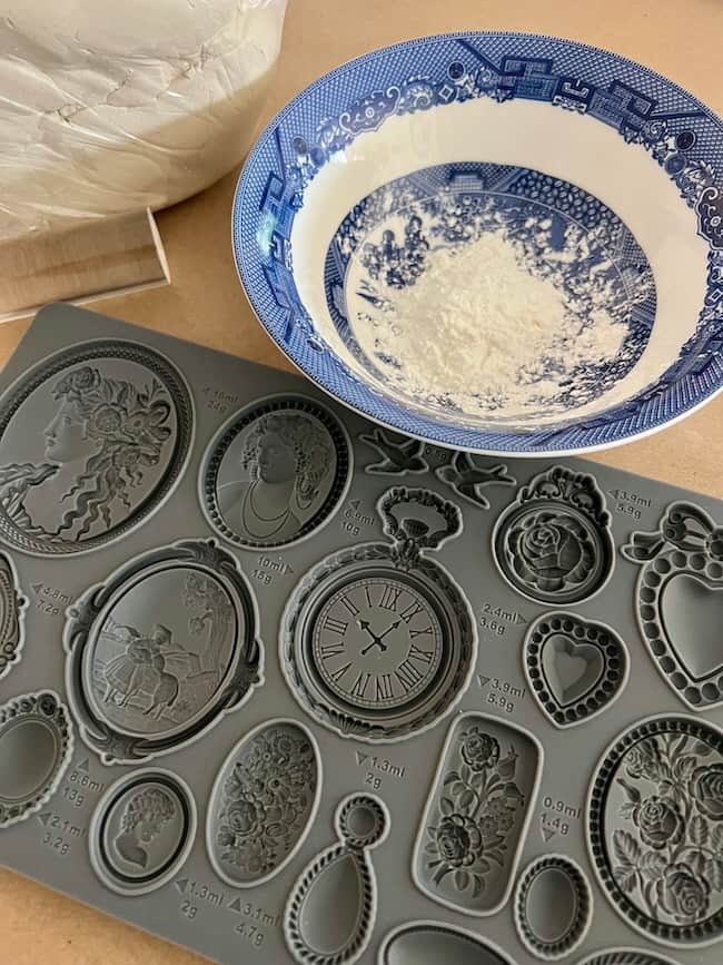 A blue and white bowl with white powder inside sits next to a gray silicone mold featuring various ornate cameo, clock, and frame designs—perfect for crafting your own DIY intaglio art projects.
