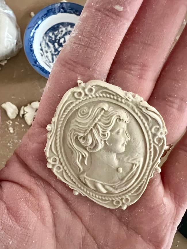 A hand holds a round, beige plaster medallion with a raised profile of a woman in an ornate frame—an inspiring example of DIY intaglio art. In the background, a small blue and white dish with paint residue hints at the creative process.