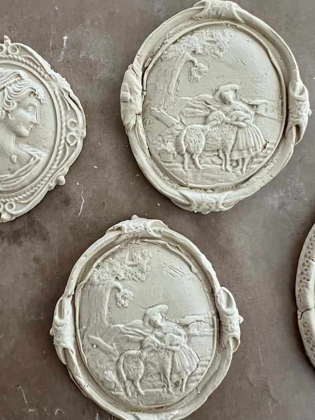 Oval plaster reliefs featuring intricate designs: one shows a woman’s profile, and two depict a pastoral scene with a woman and sheep under trees, all set against a brown background—perfect inspiration for your own DIY intaglio art.