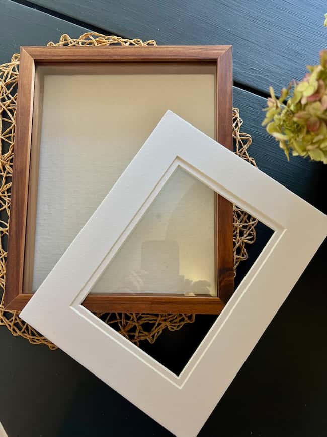 A wooden picture frame lies on a woven mat, with a white photo mat propped diagonally across it. On the right, part of a flower arrangement appears, evoking inspiration for your next DIY intaglio art project.
