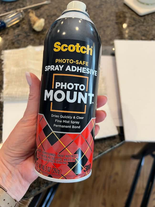 A hand holds a can of Scotch Photo Mount spray adhesive in a kitchen, with a table and papers in the background—perfect for DIY intaglio art projects requiring a permanent bond. The black can features red plaid design and bold text.