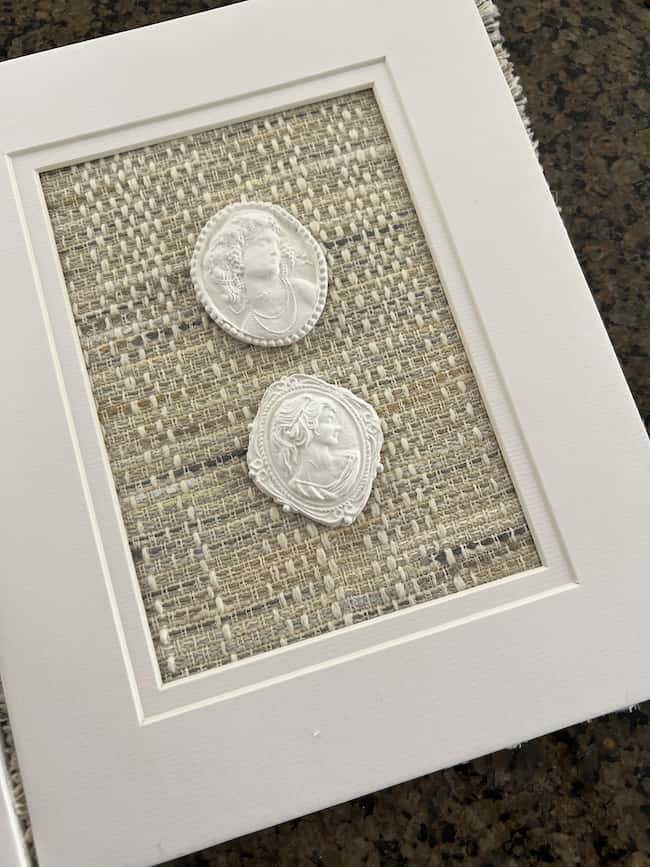 A framed display with two white wax seals featuring classical-style portraits, crafted in a DIY intaglio art style and mounted on a textured beige fabric background.