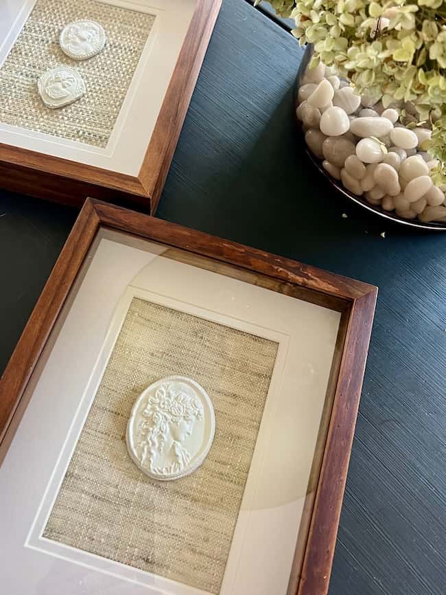 Framed white cameo and DIY intaglio art pieces are displayed on textured fabric backgrounds, set on a dark surface next to a glass bowl filled with white stones and light green dried flowers.