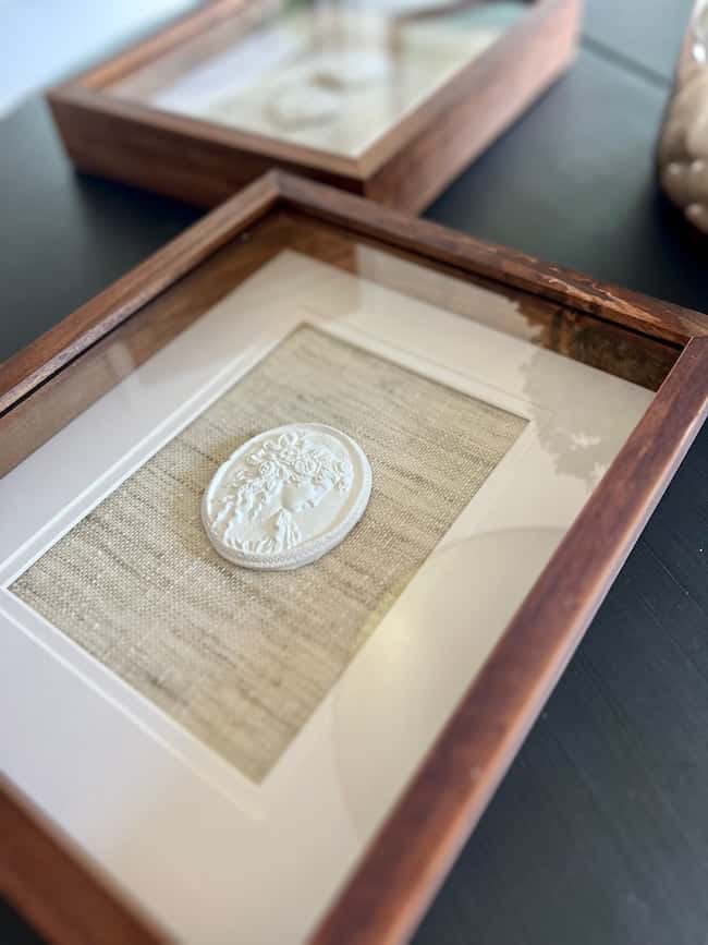 A close-up of a framed piece of DIY intaglio art featuring a white, oval-shaped embossed medallion on textured fabric, set in a wooden frame and displayed on a dark surface. Another similar frame is visible in the background.