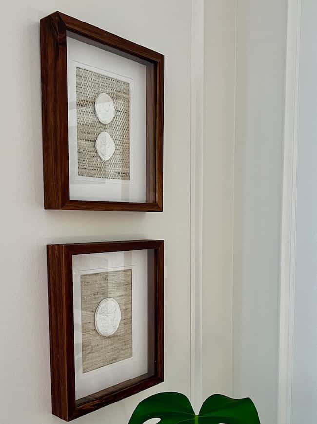 Two wooden frames hang vertically on a light-colored wall, each showcasing DIY intaglio art with white circular objects on a textured, neutral background. A green leaf is partially visible at the bottom right corner.