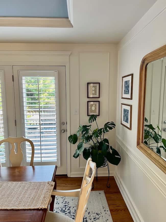 Bright dining room corner with a wooden table and chairs, a woven placemat, a large wall mirror, DIY intaglio art framed on the wall, a green potted plant, and glass doors with white shutters letting in natural light.