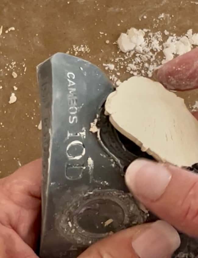 Close-up of hands removing a broken, circular makeup powder from a dark compact case. Powder pieces and dust are scattered on a brown surface underneath, ready to be repurposed for DIY intaglio art projects.