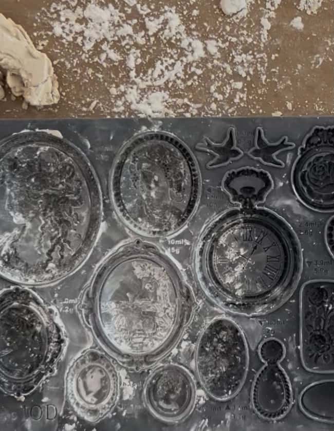 A silicone mold with various ornate cameo and clock designs, perfect for DIY intaglio art, is dusted with white powder—likely flour or cornstarch—on a messy surface, with some dough or clay in the corner.