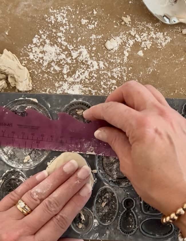 Hands pressing dough into a baking mold while using a purple ruler to trim the edges, resembling the precision of DIY intaglio art, with flour and dough scattered on a brown surface.