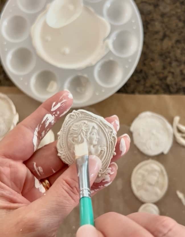 A person uses a paintbrush to apply white paint to a small, round decorative cameo featuring a woman's profile, creating unique DIY intaglio art, with a paint palette and more cameos visible in the background.