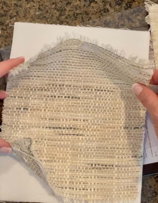 A person holds a piece of loosely woven, beige fabric with frayed edges over a white rectangular surface on a granite countertop, preparing materials for a DIY intaglio art project.