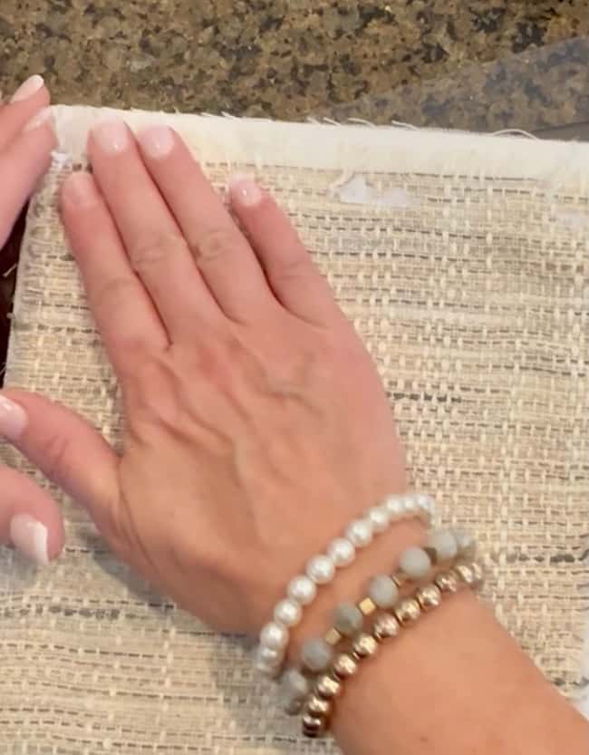 A hand with pale pink nails and several beaded bracelets smooths out a piece of woven, beige fabric on a speckled countertop—perfect for inspiring your next DIY intaglio art project.