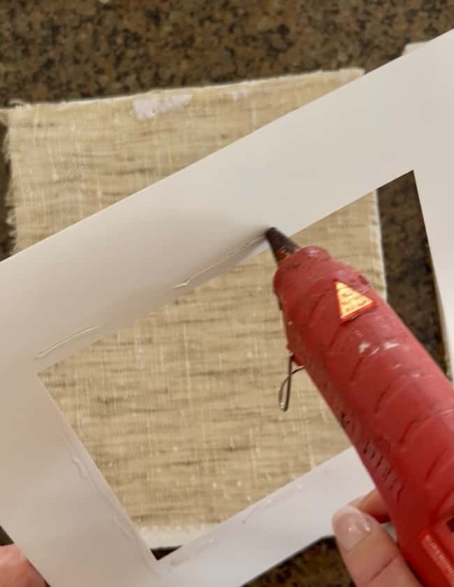 A hand holds a red hot glue gun, applying glue to the edge of a white rectangular frame—perfect for preparing a base for DIY intaglio art—with coarse beige fabric lying on a dark surface in the background.