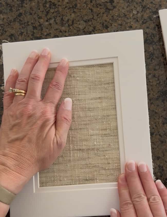 Two hands placing a piece of textured beige fabric into a white photo frame on a dark, speckled surface, creating unique DIY intaglio art. One hand wears a wedding ring and a light-colored watch.