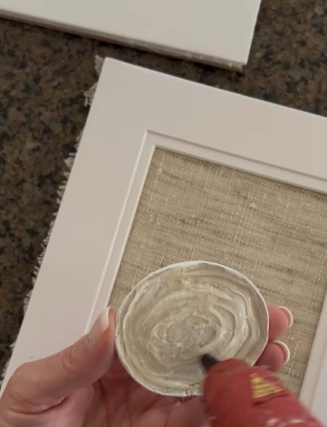 A hand holds a round seashell being glued with a hot glue gun for a DIY intaglio art project, with a white picture frame and textured beige background visible underneath.