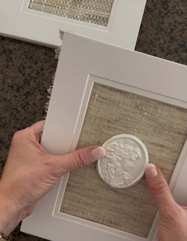 Two hands are holding a white picture frame with textured beige fabric and a decorative white medallion, showcasing DIY intaglio art, while another similar frame rests nearby on a dark surface.