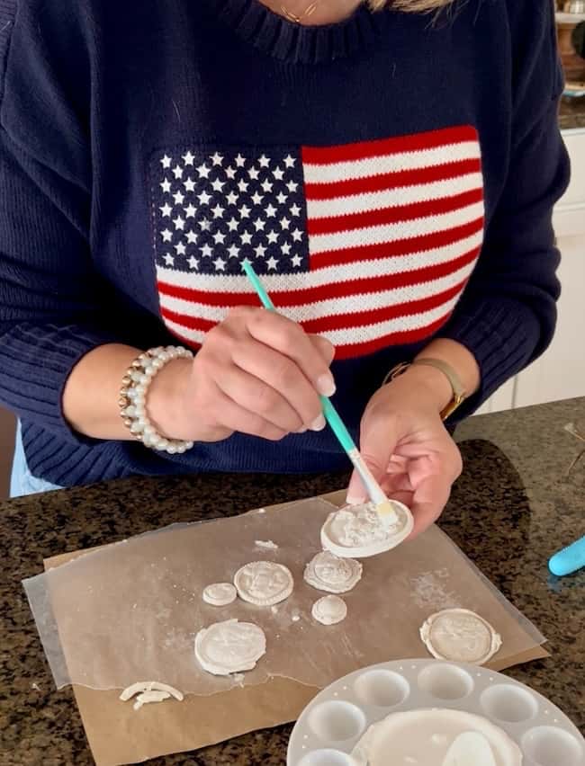 A person wearing a navy sweater with an American flag design creates DIY intaglio art, painting white clay medallions on parchment paper, with art supplies visible on the countertop.