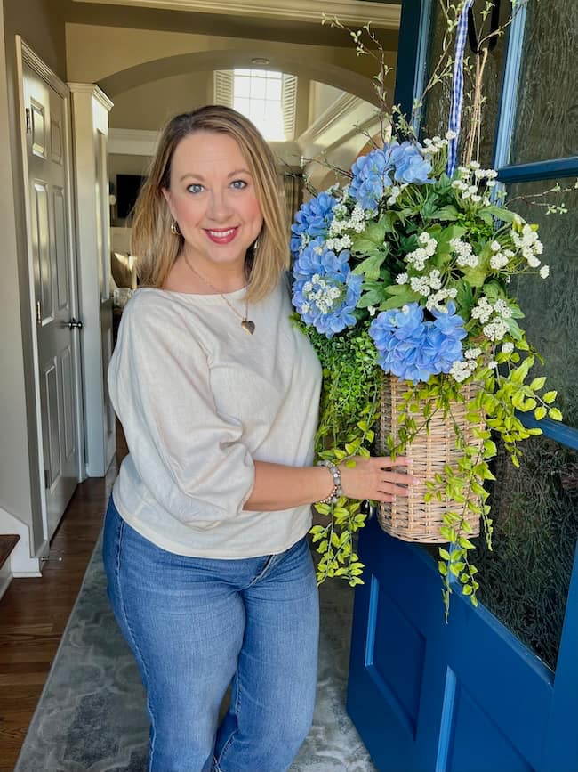 A woman with blonde hair, wearing a light gray top and jeans, stands smiling at an open blue door while holding a large basket of blue and white flowers with greenery. Indoor hallway and natural light are visible behind her.