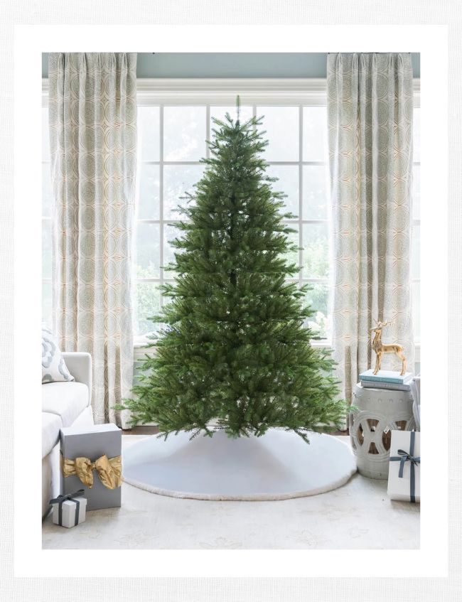 A decorated Christmas tree, fluffed and lit to perfection, stands in front of large windows with patterned curtains. Wrapped gifts, including one with a gold ribbon and a small gold reindeer figurine, rest nearby on the floor and a white side table.