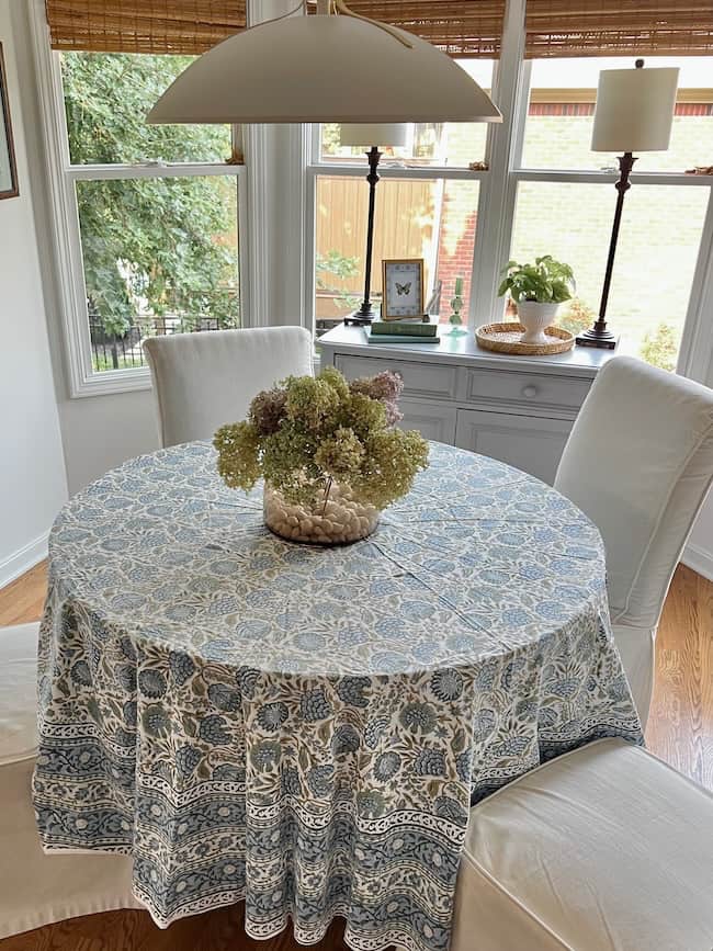 A round table with a blue and white patterned tablecloth is centered in a bright room with large windows, creating a Cozy Start to the season. The table holds a vase of dried flowers and is surrounded by two white upholstered chairs.