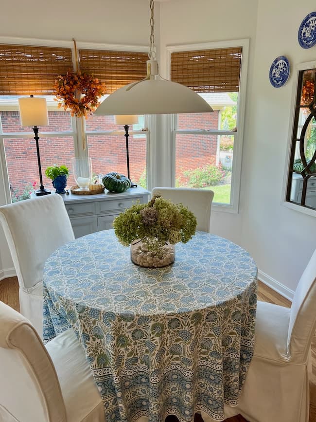 A cozy round table with a blue and white patterned tablecloth and a vase of dried flowers sits in a bright dining nook, perfect for a simple fall home tour, with four white chairs and large windows showing a brick wall outside.