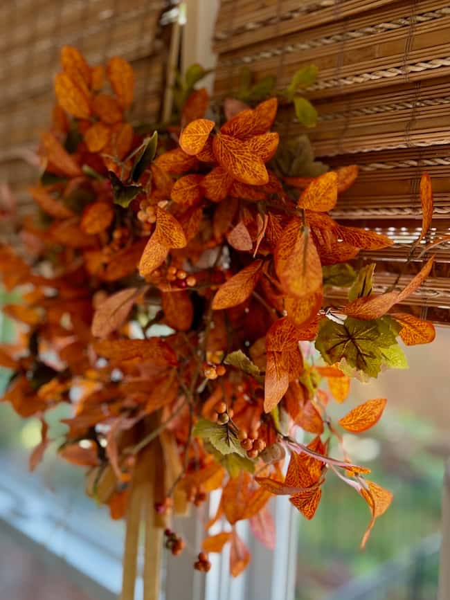 A wreath of orange and green autumn leaves hangs on a window with bamboo blinds, adding a cozy touch to your simple fall home tour decor.
