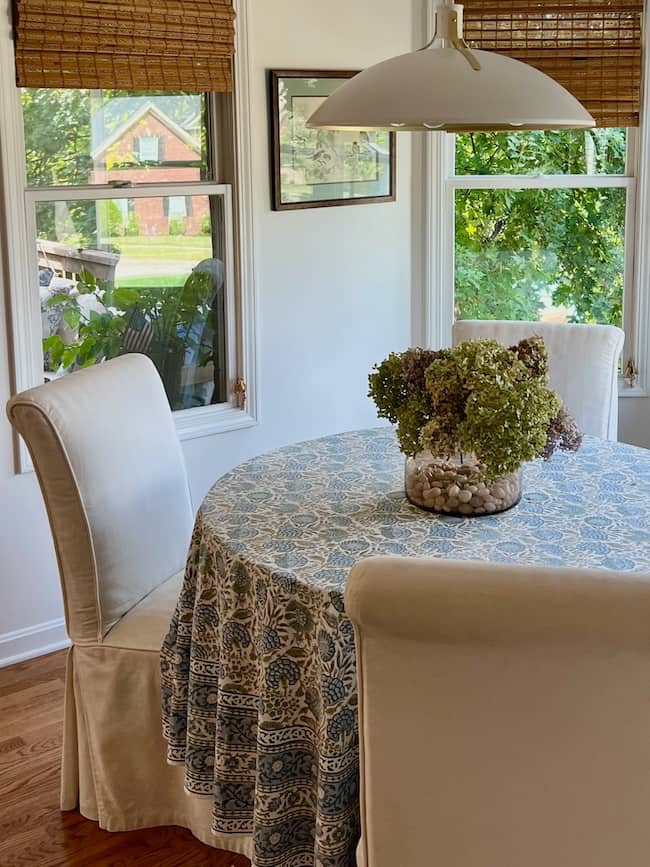 A round table with a blue-patterned tablecloth and a glass vase filled with dried flowers sits by three large windows, surrounded by white upholstered chairs in a bright, cozy dining area—perfect for a simple fall home tour.