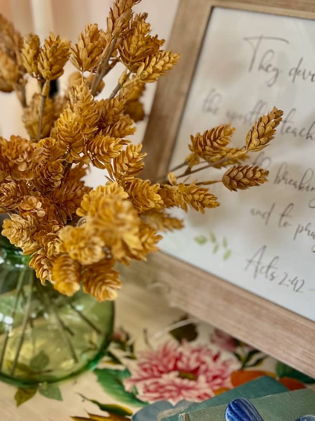 A green glass vase holds dried yellow wheat stems on a floral tablecloth beside a framed print with handwritten Acts 2:42—a cozy simple fall home tour scene.