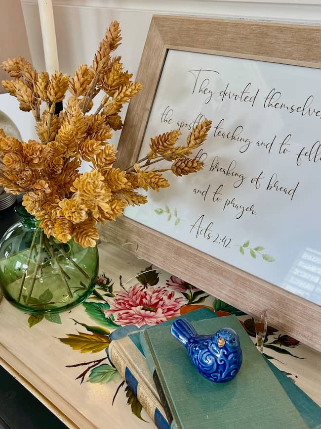 A vase of dried yellow flowers sits on a floral-patterned table next to two books, a small blue bird figurine, and a framed Bible verse—perfect touches for a cozy simple fall home tour.