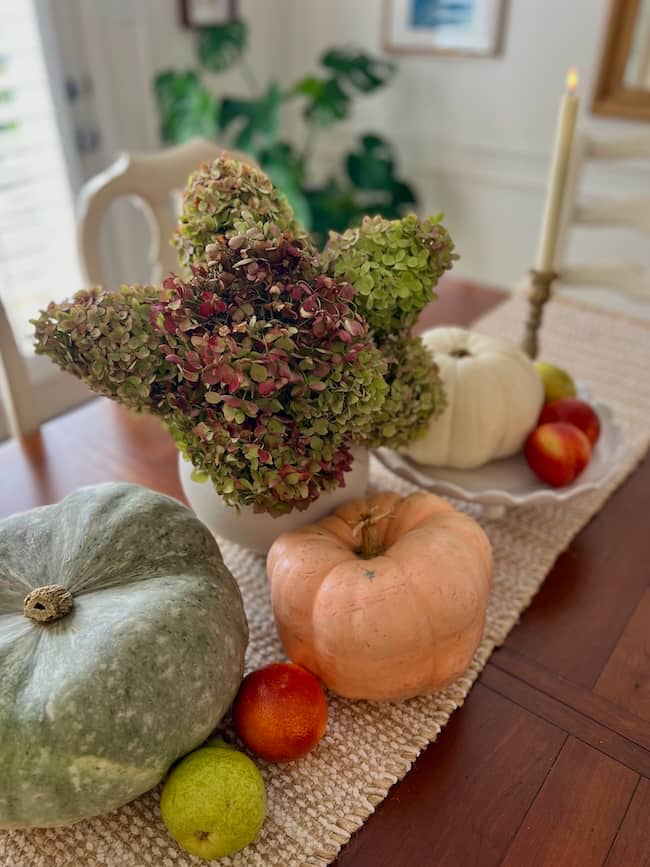 A cozy simple fall home tour centerpiece featuring pumpkins, apples, and a green pear on a woven table runner, with a vase of dried hydrangeas and a lit taper candle in the background.