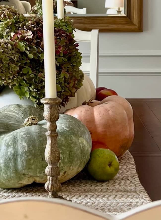 A rustic dining table centerpiece sets the scene for a cozy simple fall home tour, featuring a tall candlestick, green and orange pumpkins, hydrangeas, apples, and a pear atop a textured runner. A framed mirror hangs on the wall in the background.
