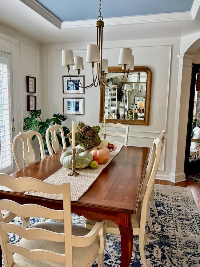 A cozy dining room with a wooden table, cream-colored chairs, a white runner, candles, and assorted gourds as a centerpiece—perfect for a simple fall home tour. A gold-framed mirror and artwork hang on the walls beneath the chandelier.