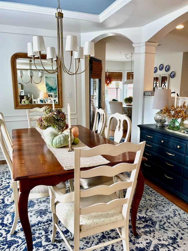 A cozy dining room with a wooden table, six white chairs, a blue-patterned rug, and a chandelier. The table is styled for a simple fall home tour with gourds and flowers. A navy sideboard and large mirror complete the inviting space.