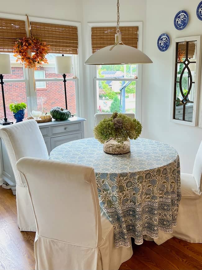 A round dining table with a blue patterned tablecloth is surrounded by four white slipcovered chairs. A large floral centerpiece sits on the table. Bright windows and autumn decor set the scene for a cozy simple fall home tour.